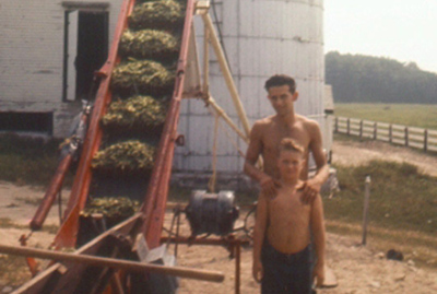 1957 – Bob Gagnon standing with Warren Shaw, Jr., loading corn into silo next to a corn elevator.